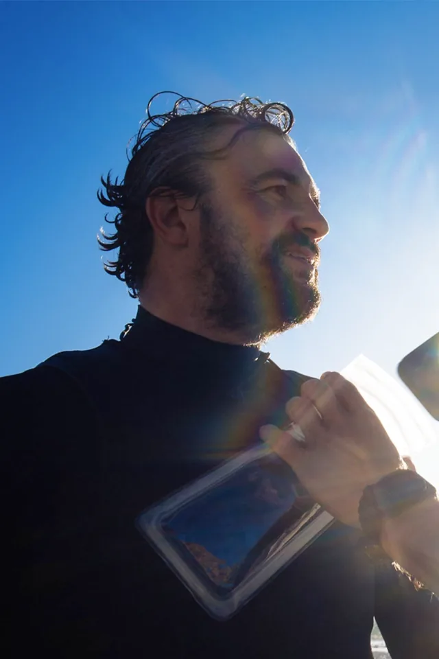 Christian Rolly, bearded and in a black wetsuit, looking out to sea in bright sunlight