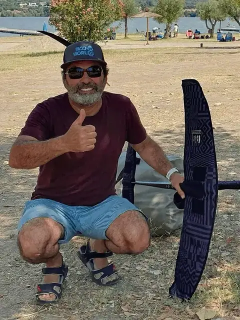 Juan Miquel, in a WOO Worlds cap, crouching beside a foil wing and giving a thumbs up at a lakeside